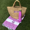 Purple 100% Cotton Bold Stripe Beach Towel with a large single letter monogram. On top is a basket bag with a large three letter monogram, and a pink bottle with a three letter monogram.  
