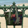 Large Camo style Tote bag, with a large neon embroidered monogram, hung on railings by a beach 